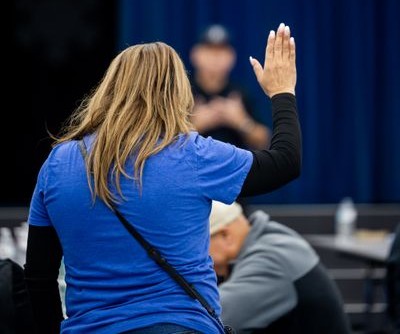 A woman in a blue shirt raises her hand in a classroom or meeting setting, engaging with the speaker.